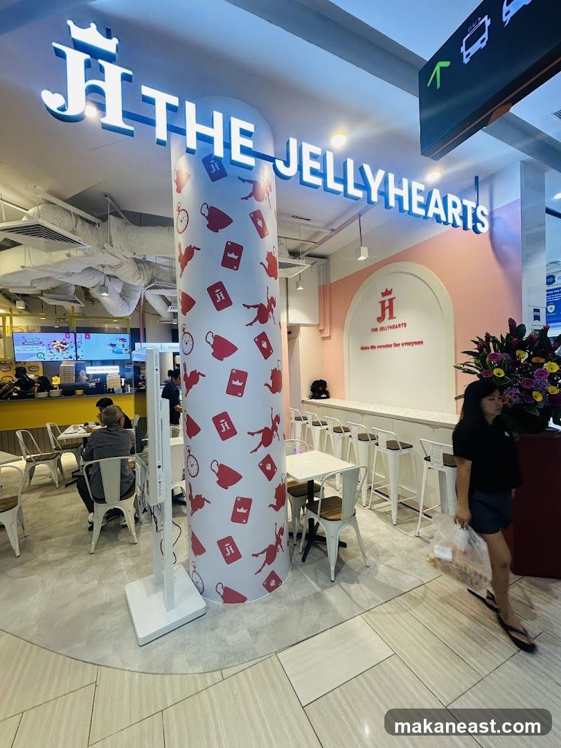A brightly lit storefront with a prominent sign reading "THE JELLYHEARTS" in large white letters. The pillar in front of the store is decorated with a whimsical pattern of teacups, playing cards, and silhouettes. Inside, there are tables and chairs, a counter with stools, and a display screen. A woman is walking past the entrance carrying a plastic bag.
