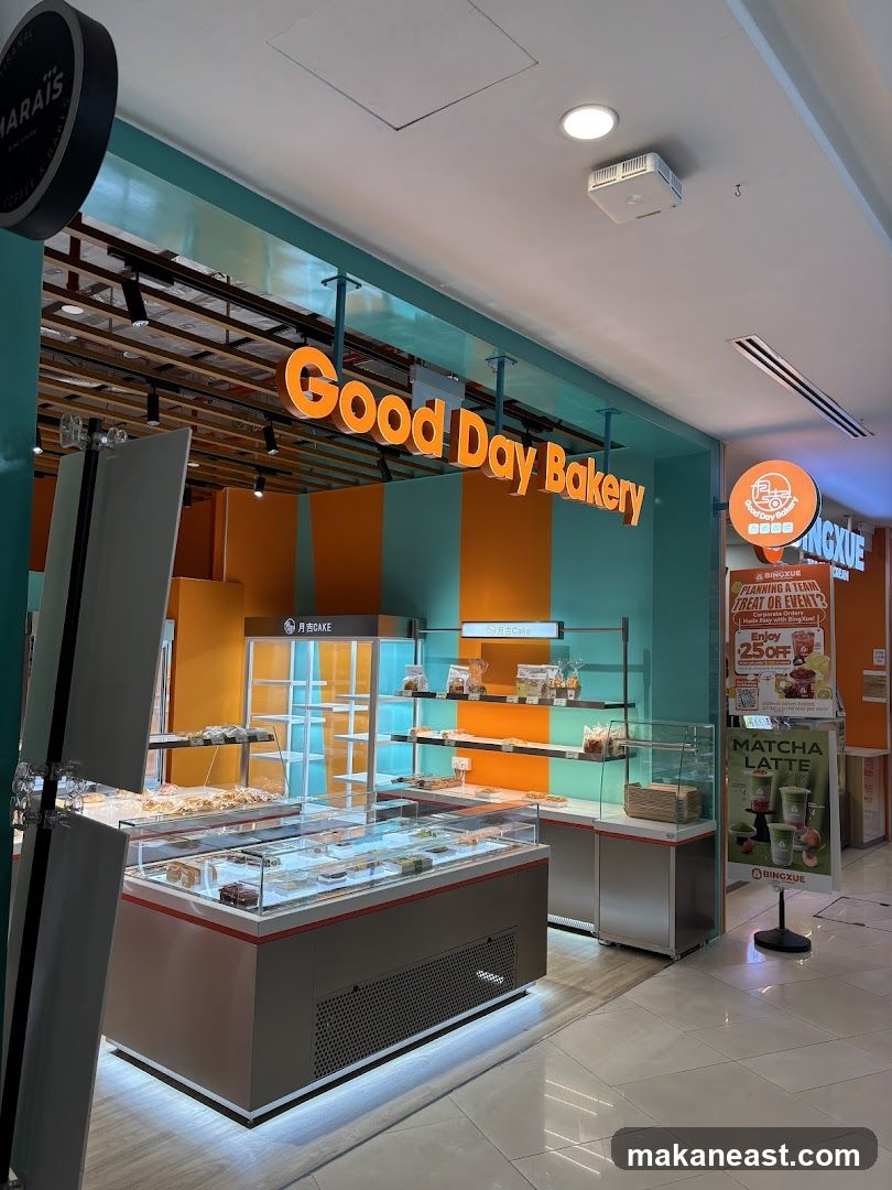 The exterior of a bakery with a prominent "Good Day Bakery" sign. Display cases filled with pastries are visible inside. The shop has a vibrant orange and teal color scheme. A promotional poster for a discount and a sign for "Matcha Latte" are also visible.