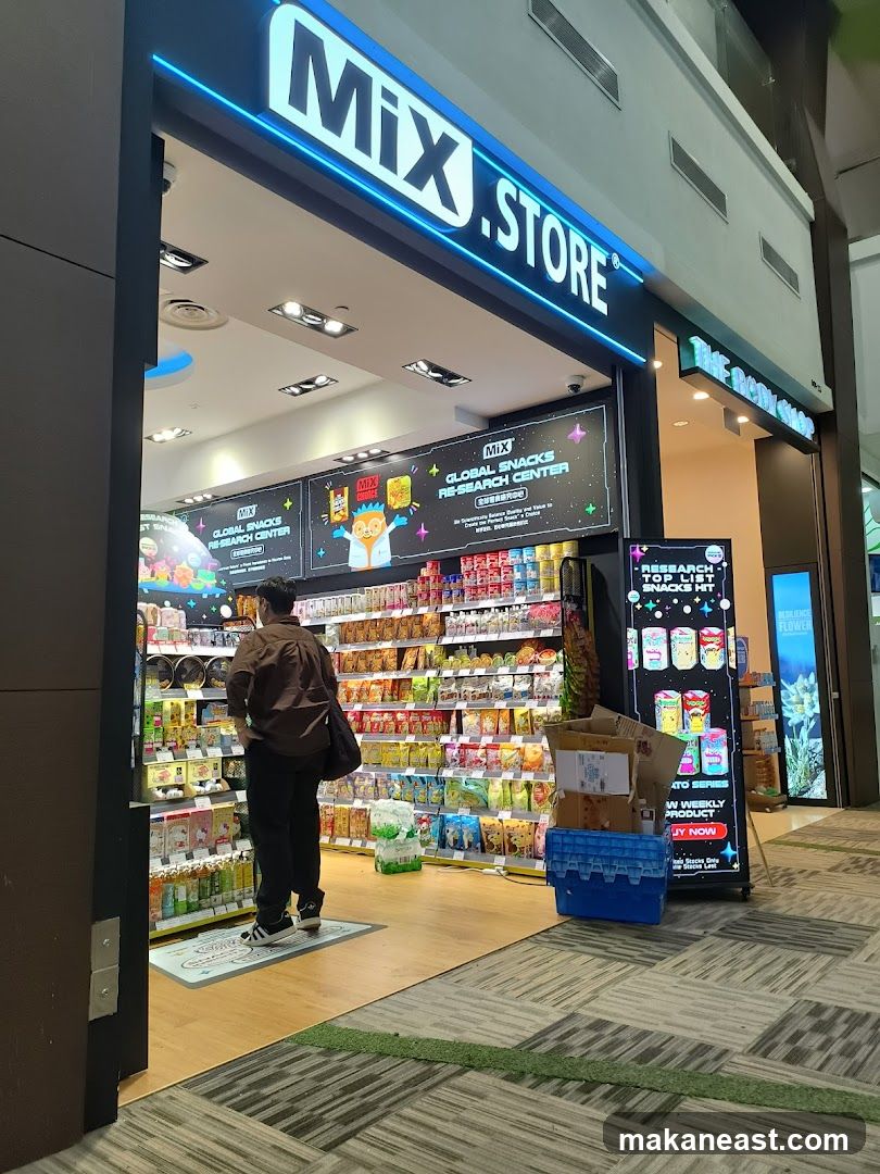 The exterior of a store called Mix Store, which sells global snacks. The store has a bright, modern facade with a large illuminated sign. Inside, shelves are stocked with various snacks and products. A person is standing in the entrance looking at the merchandise.