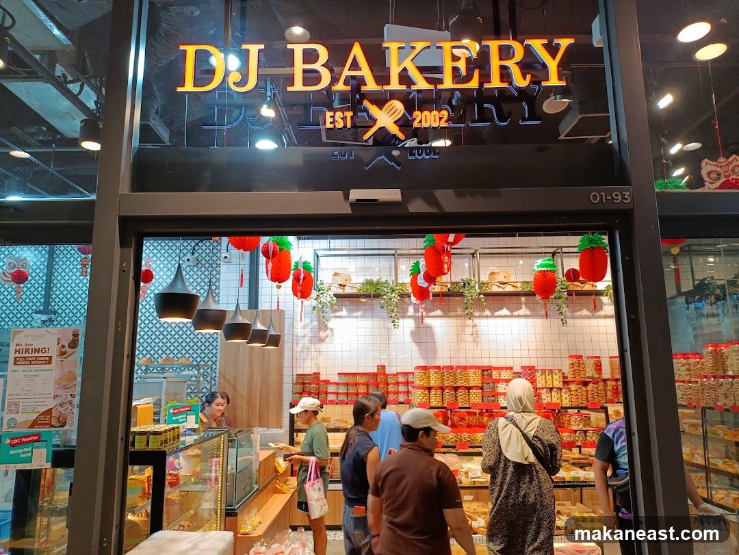 The exterior of DJ Bakery, a shop selling baked goods. The shop has a large orange sign with the name "DJ BAKERY EST 2002" above the entrance. Inside, shelves are stocked with various packaged snacks and pastries. Several customers are browsing and purchasing items. Red and green decorative fruit-shaped lanterns hang from the ceiling.