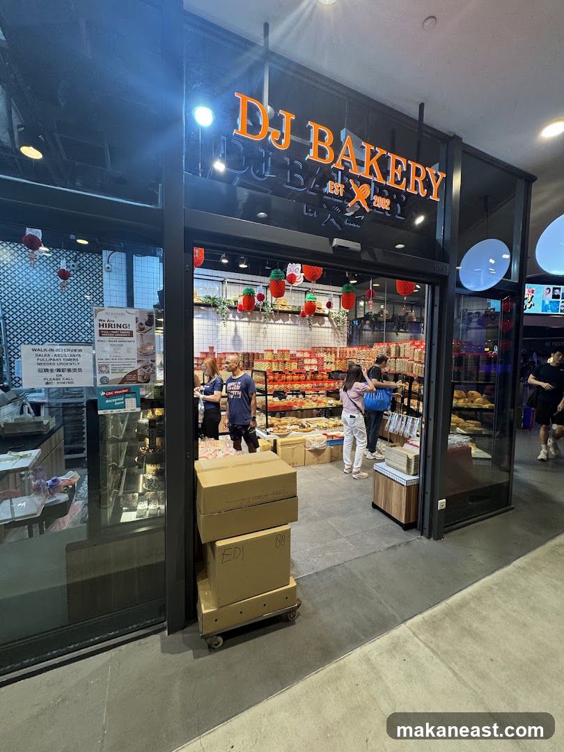 The exterior of a bakery with a large sign that reads "DJ BAKERY EST 2002". Inside, shelves are stocked with various baked goods and packaged items. Several people are visible inside the shop, and there are decorative strawberry-shaped ornaments hanging from the ceiling. Boxes are stacked on a trolley outside the entrance. A "HIRING!" poster is displayed on the left side of the entrance.