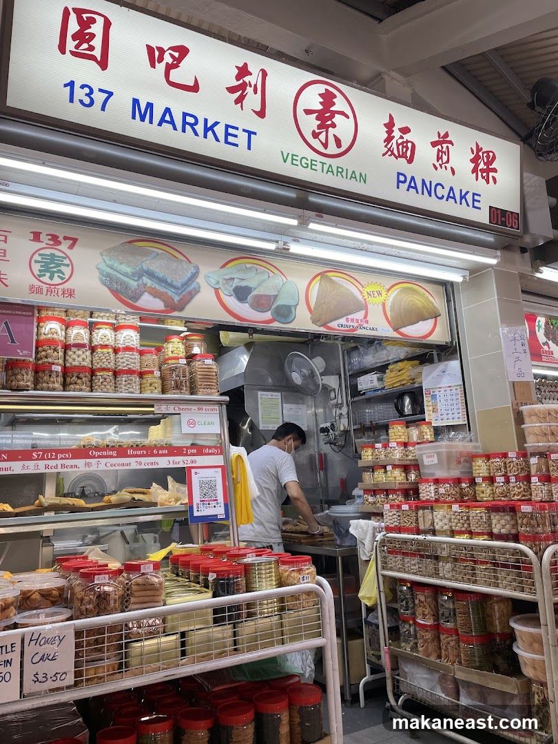 A street-level view of a food stall with a prominent sign advertising "137 MARKET VEGETARIAN PANCAKE". The stall displays a variety of snacks and food items in jars and containers, with images of pancakes and other dishes shown above the counter. A person in a grey t-shirt and mask is visible working behind the counter.