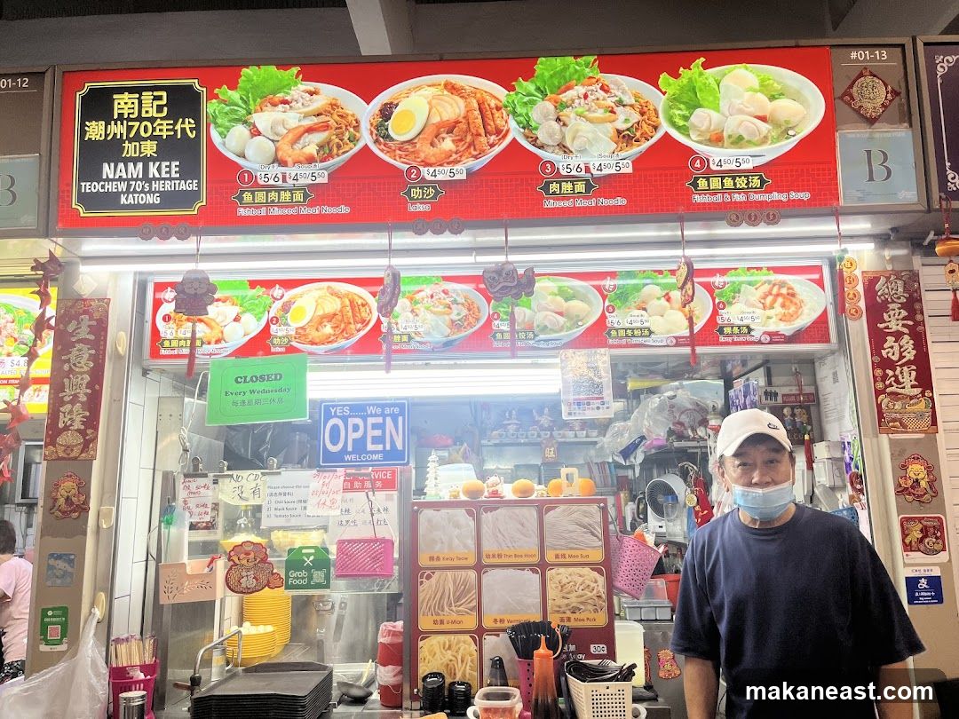 A food stall with a large red sign displaying various noodle dishes and their prices. Below the sign, there is a counter with ingredients and a man wearing a cap and mask. A green sign indicates the stall is closed every Wednesday.