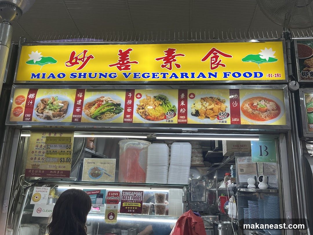 A brightly lit yellow sign with Chinese characters and the English text "MIAO SHUNG VEGETARIAN FOOD" is displayed above a food stall. Below the main sign, there are images of various dishes with their names and calorie counts. A menu board with prices is visible below the images. The stall appears to be in a hawker center, with stacks of containers and plates visible.