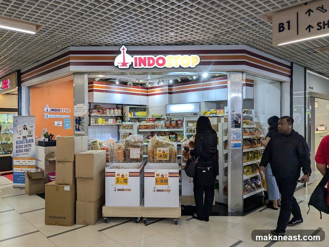 A view of the Indostop store entrance in a mall. The store has a prominent "INDOSTOP" sign above the entrance. Shelves stocked with various products are visible inside. Several boxes are stacked outside the store. People are seen walking by the storefront.