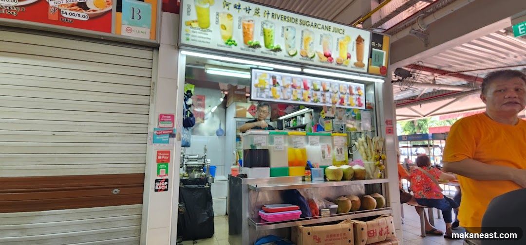 A sugarcane juice stall with a menu board displaying various drinks and prices. Coconuts and sugarcane stalks are visible. A man is seen preparing drinks behind the counter.