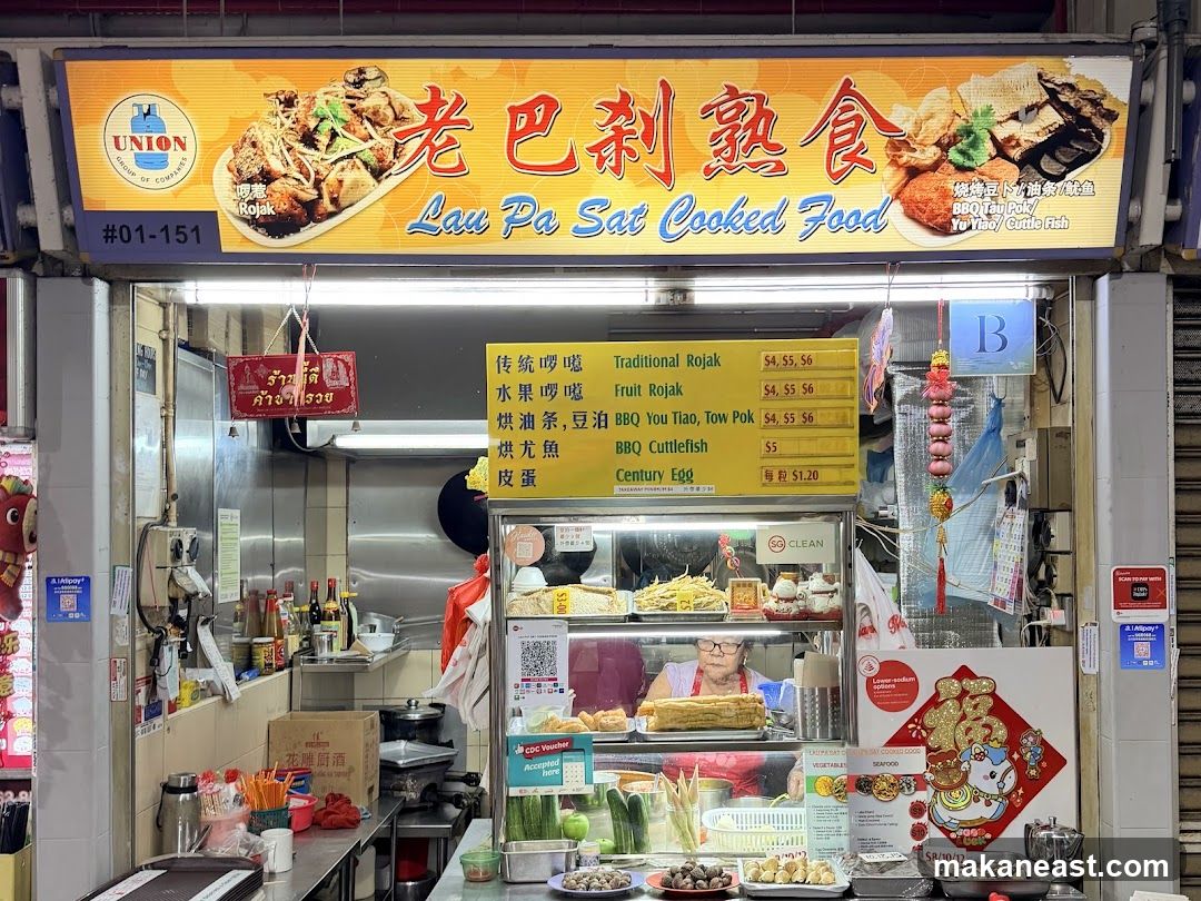 A food stall with a large yellow sign featuring Chinese characters and English text "Lau Pa Sat Cooked Food". The sign also includes images of food and a logo for "UNION GROUP OF COMPANIES". Below the main sign is a yellow menu board listing various dishes and prices. The stall displays food items in glass cases, and a person is visible behind the counter. Various decorations and signage are present, including a red sign with Thai script and a "SG CLEAN" certification. The stall number "#01-151" is visible.