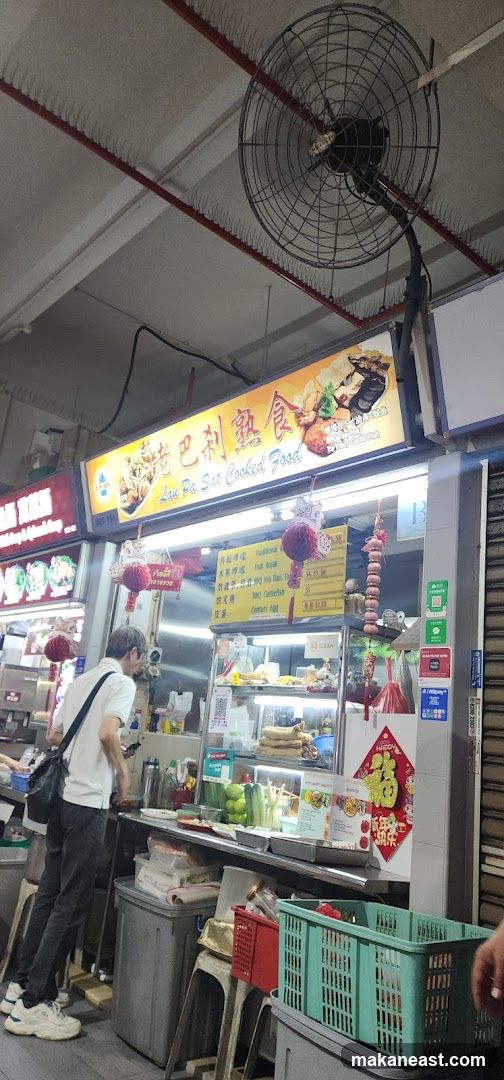 A food stall with a bright yellow sign displaying Chinese characters and the text "Lau Pa Sat Cooked Food". The stall has a display case with food items and a menu board. A man is standing in front of the stall, looking at his phone. There are decorative red lanterns hanging from the stall.
