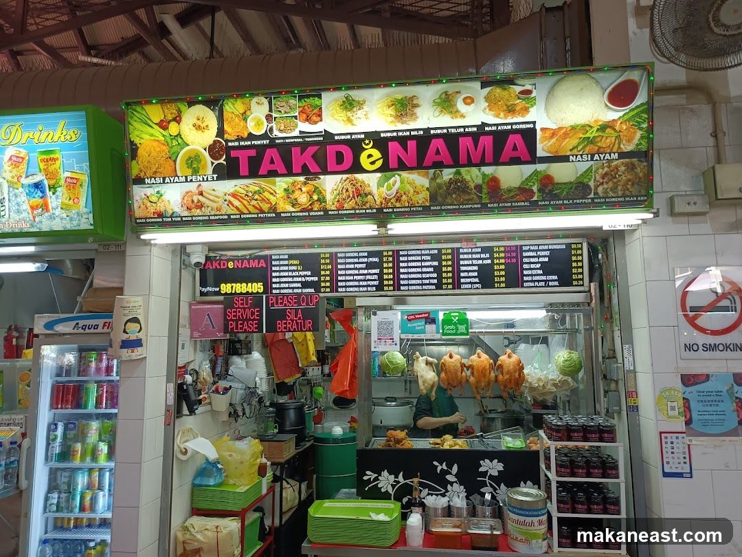 A food stall with a large illuminated sign displaying various dishes and the name 'TAKDENAMA'. Below the sign is a menu board with prices and a display of food items including roasted chickens. The stall also has refrigerators stocked with drinks and shelves with jars of condiments.