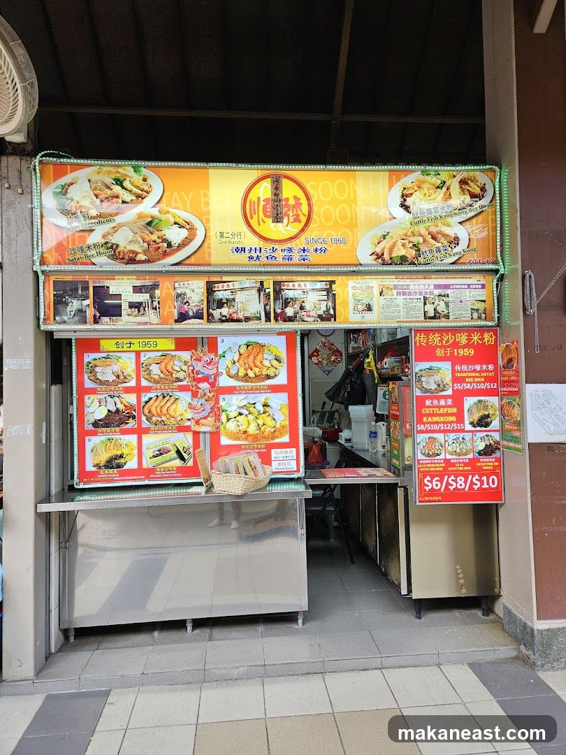 A food stall with a large illuminated sign displaying food images and prices. The stall has a metal counter and various menu boards.
