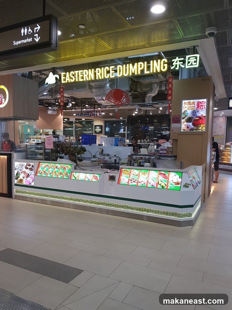 A brightly lit food stall with a prominent sign reading "EASTERN RICE DUMPLING" in English and Chinese characters. The stall features a display counter showcasing various food items, likely dumplings and rice dishes, presented in illuminated panels. Behind the counter, staff members are visible preparing food. The stall is situated in a mall or food court, with a directional sign indicating "Supermarket" nearby.