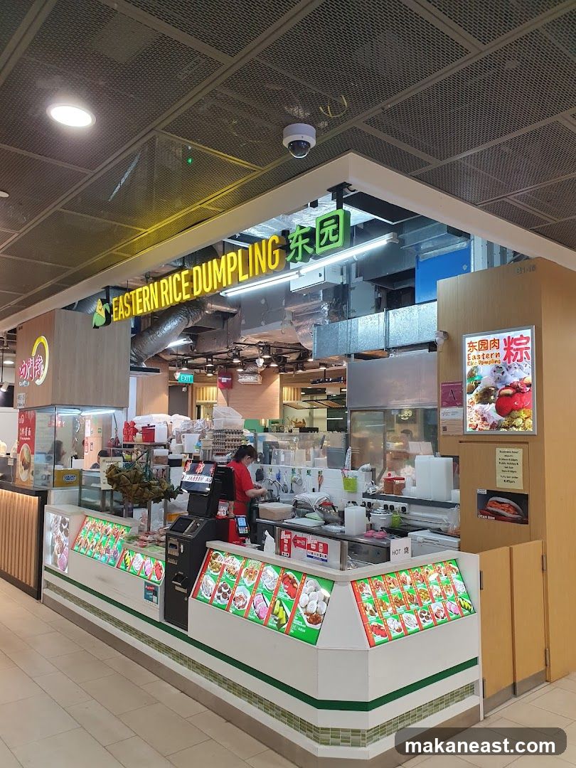 A food stall with a prominent sign that reads "EASTERN RICE DUMPLING" in English and Chinese characters. The stall displays various food items in refrigerated cases, with images of dumplings and other dishes. A staff member is visible behind the counter.