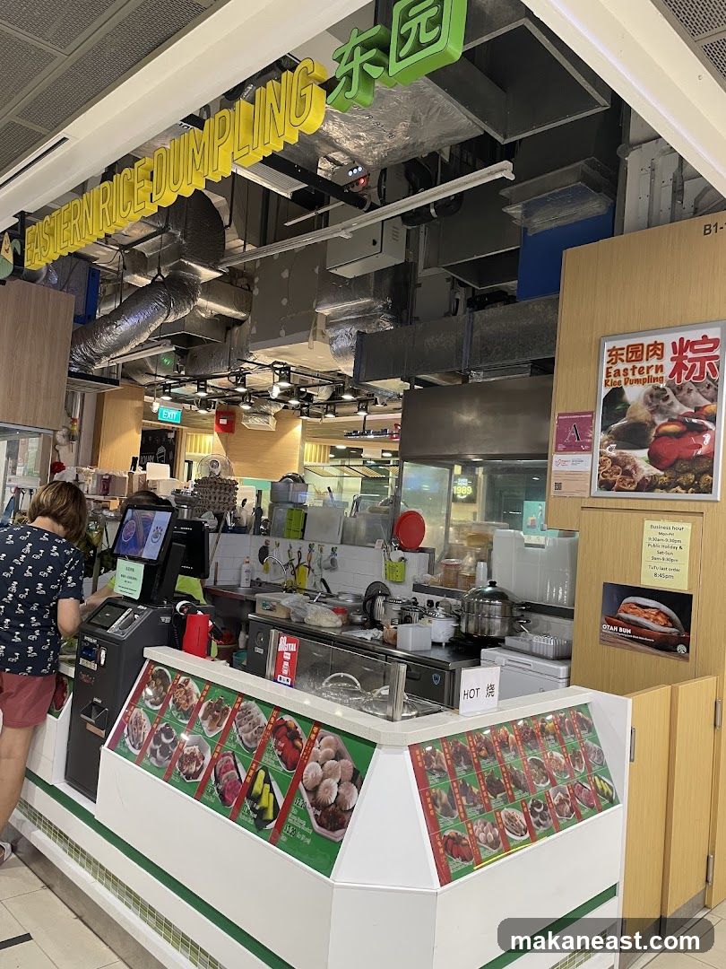 A food stall with a prominent yellow sign reading "EASTERN RICE DUMPLING" and green Chinese characters. The stall displays various food items in a refrigerated case with pictures and prices. A person is interacting with a self-service kiosk. Behind the counter, kitchen equipment and supplies are visible. A poster advertises "OTAH BUN" and lists business hours.