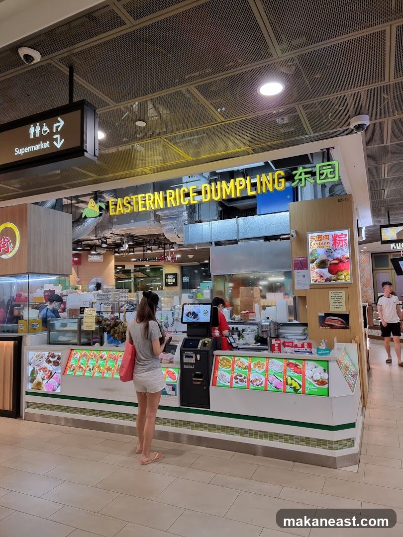 A food stall named Eastern Rice Dumpling with a display of various food items. A woman is standing in front of the counter, looking at a menu or ordering. There are employees behind the counter preparing food. The stall is located in a mall or food court.