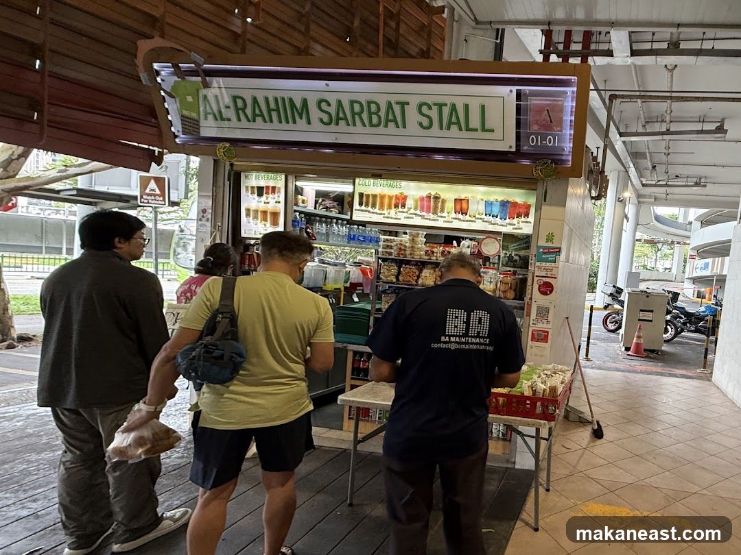 A view of the Al-Rahim Sarbat Stall storefront with customers. The stall has a sign that reads "AL-RAHIM SARBAT STALL" and displays various hot and cold beverages.