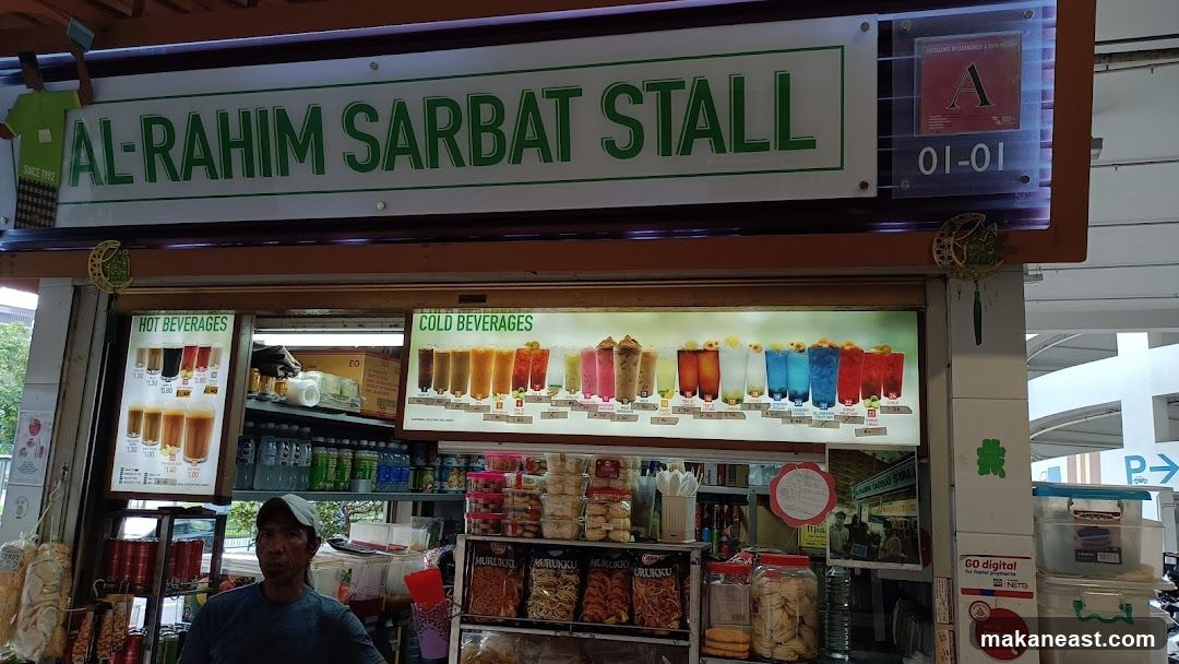 The exterior of a food stall with a prominent sign that reads "AL-RAHIM SARBAT STALL". Below the sign, illuminated displays showcase "HOT BEVERAGES" and "COLD BEVERAGES" with images of drinks and prices. Shelves behind the counter are stocked with various bottled drinks and packaged snacks. A man wearing a cap is visible behind the counter.