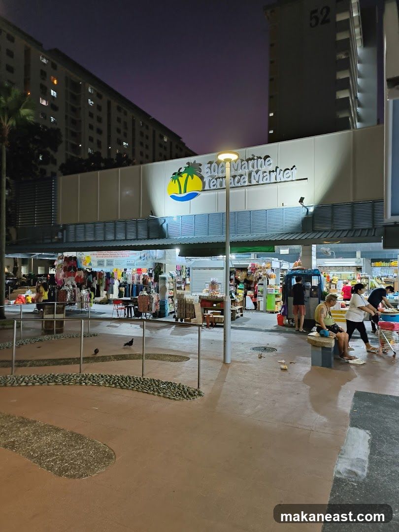 The exterior of the 50A Marine Terrace Market at dusk. The market has a white facade with a prominent sign featuring a tropical island logo and the text "50A Marine Terrace Market". Below the sign, there are multiple stalls selling various goods, including clothing and food items. People are seen browsing and shopping.