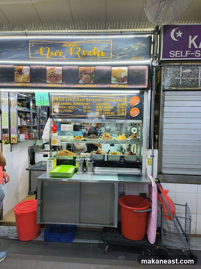 A food stall with a sign displaying the name "Aur Rezki" and pictures of various dishes. Below the name, a price list is visible. The stall has a display case with food items and a counter for service.