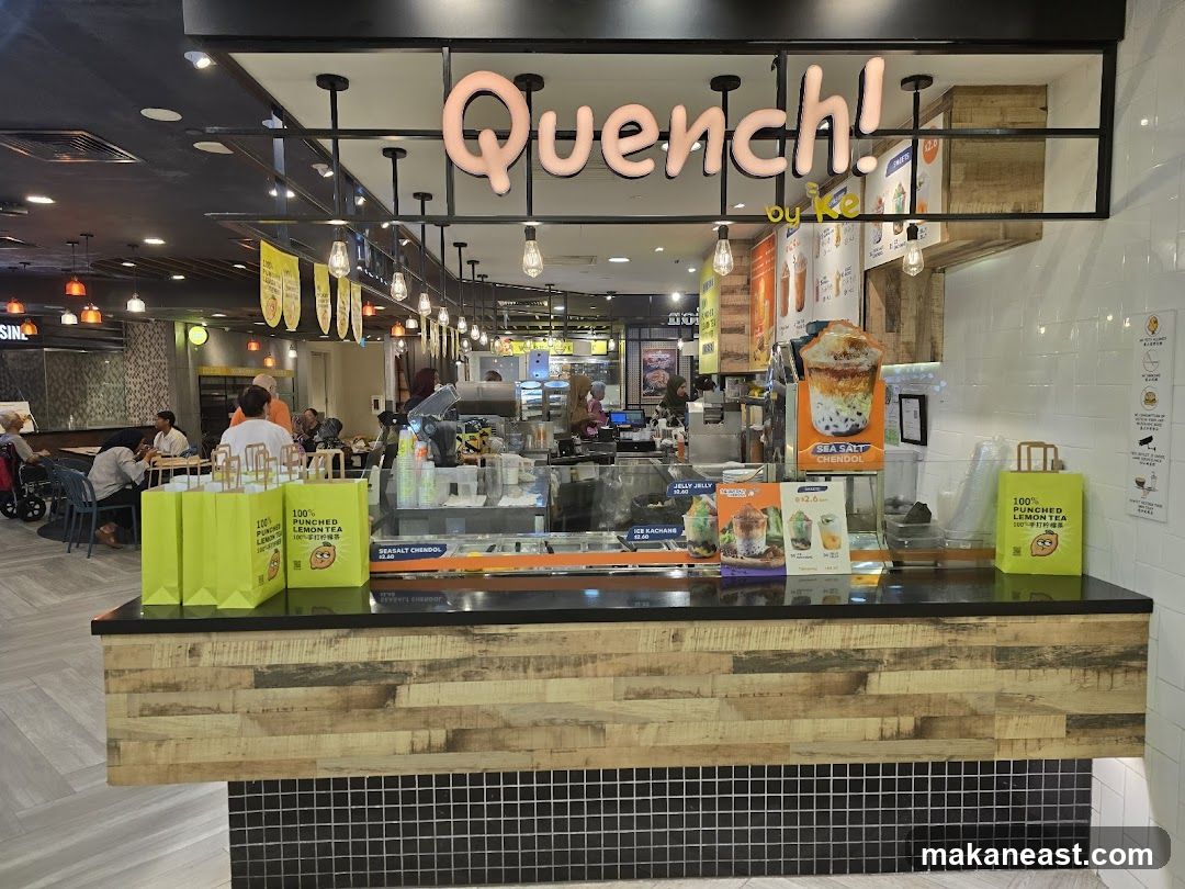 A brightly lit bubble tea shop counter with a wooden facade. The shop's name, "Quench! by Ke," is prominently displayed in large, pink, bubbly letters above the counter. Several yellow shopping bags with the shop's logo and text "100% PUNCHED LEMON TEA" are arranged on the counter. Behind the counter, various drinks and menu items are visible, including a large promotional image of a layered drink labeled "SEA SALT CHENDOL." The interior of the shop and seating area are visible in the background.