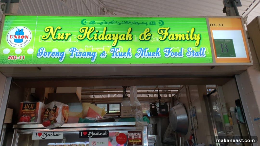A brightly lit sign for "Nur Hidayah & Family" food stall, specializing in "Goreng Pisang & Kueh Mueh". The sign features the Union Group of Companies logo and the stall number #01-11. Below the sign, the entrance to the stall is visible, with various food items and containers on display.