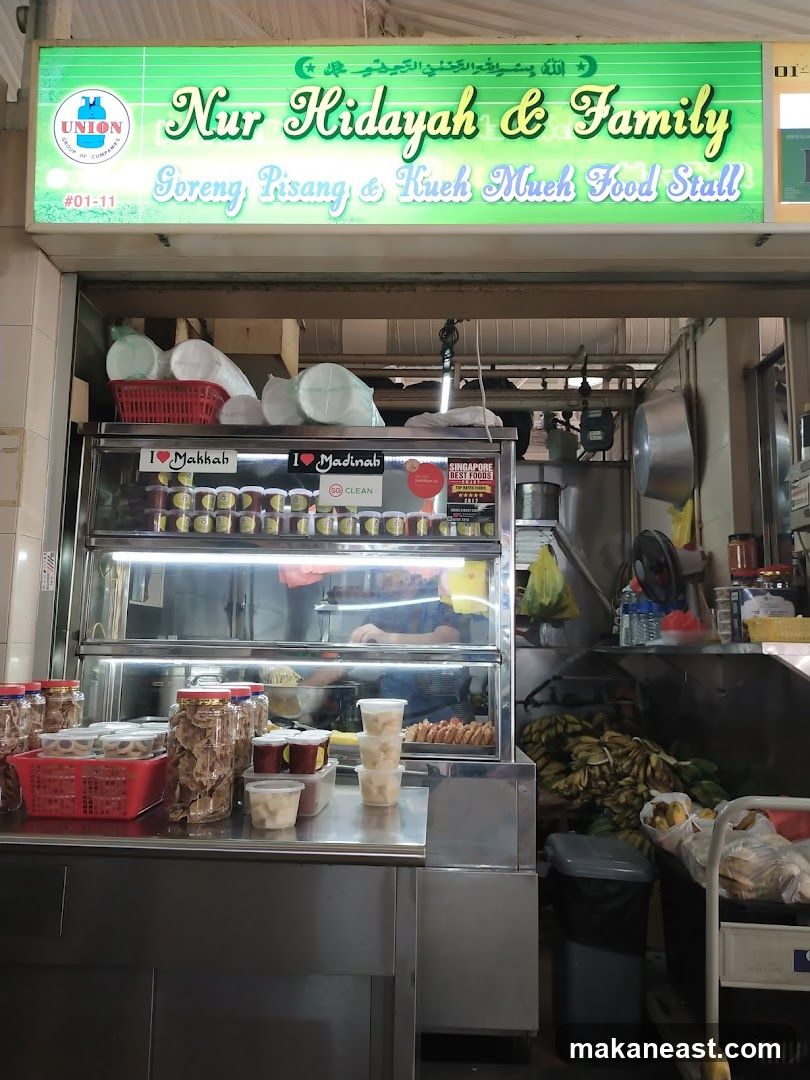 A food stall with a green illuminated sign that reads "Nur Hidayah & Family Goreng Pisang & Kueh Much Food Stall" and a "UNION GROUP OF COMPANIES" logo. Below the sign, a display case shows various food items in containers and jars. There are also bananas and other produce visible.