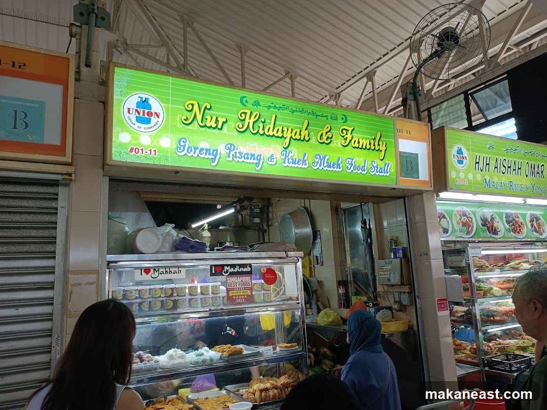 A food stall with a bright green sign that reads "Nur Hidayah & Family" and "Goreng Pisang & Kueh Much Food Stall". Below the sign, a display case shows various food items. To the right, another stall is visible with a sign for "HJH Aishah Omar Malay Rice & Yong".