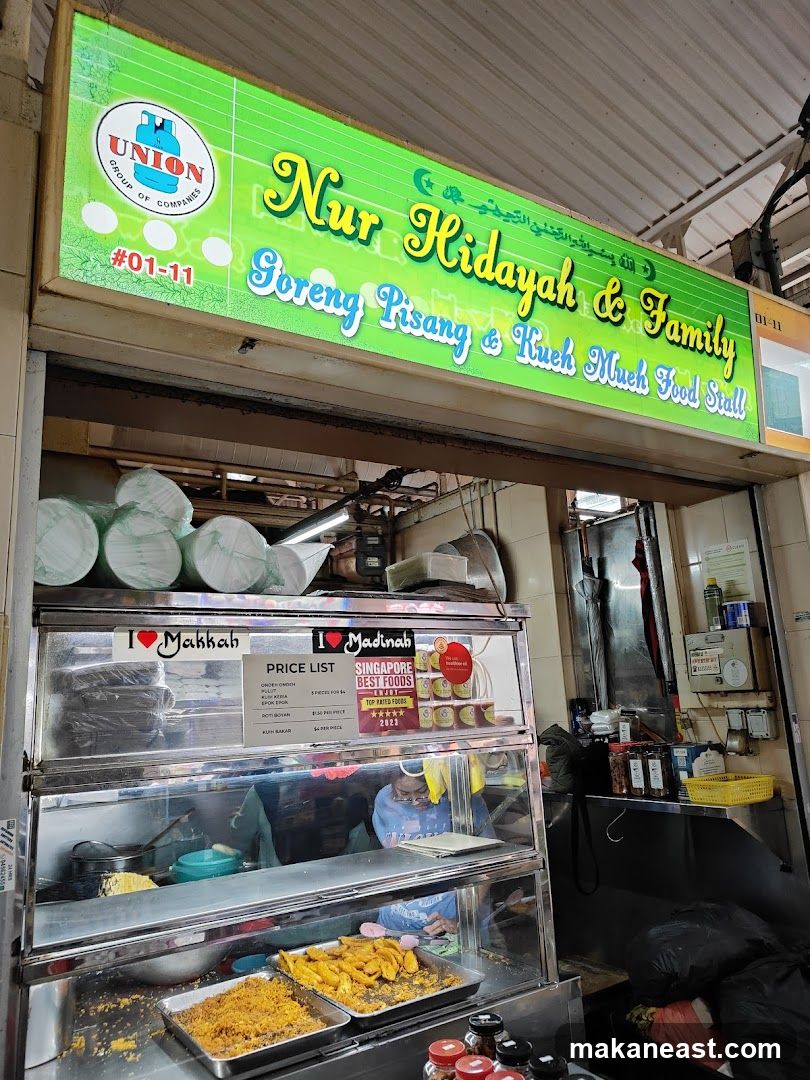 A food stall with a green illuminated sign that reads "Nur Hidayah & Family Goreng Pisang & Kueh Much Food Stall" and a "UNION GROUP OF COMPANIES" logo. Below the sign is a display case with food items, including fried bananas and other snacks. A price list is visible inside the display case.