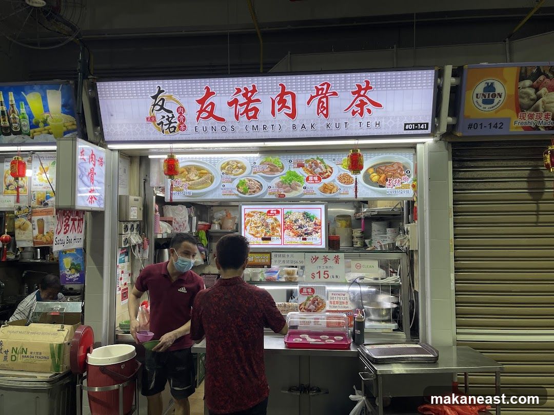 A food stall with a large illuminated sign displaying the name "Eunos (MRT) Bak Kut Teh" in English and Chinese. The sign also features images of various dishes. Two men are visible at the counter, one wearing a mask and holding bowls. Various food items and drinks are displayed on posters and within the stall.
