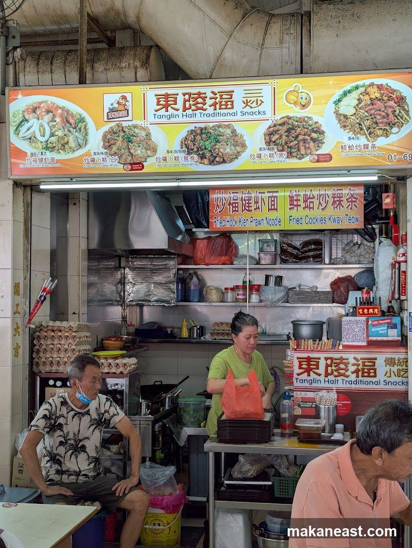 A food stall with a large overhead sign displaying images and names of various dishes. Below the sign, staff are visible preparing food. The stall appears to be in an open-air market or food court setting.
