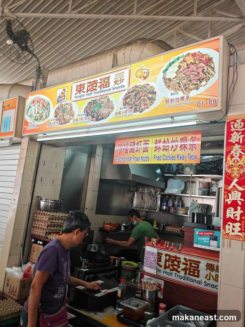 A food stall with a large illuminated sign displaying pictures of various dishes and their prices. The sign reads "Tanglin Halt Traditional Snacks" in English and Chinese. Below the main sign, another banner lists "Fried Hock Kien Prawn Noodle" and "Fried Cookies Kway Teow". Two staff members are visible inside the stall, one preparing food and the other attending to a customer. There are stacks of eggs and various ingredients visible.
