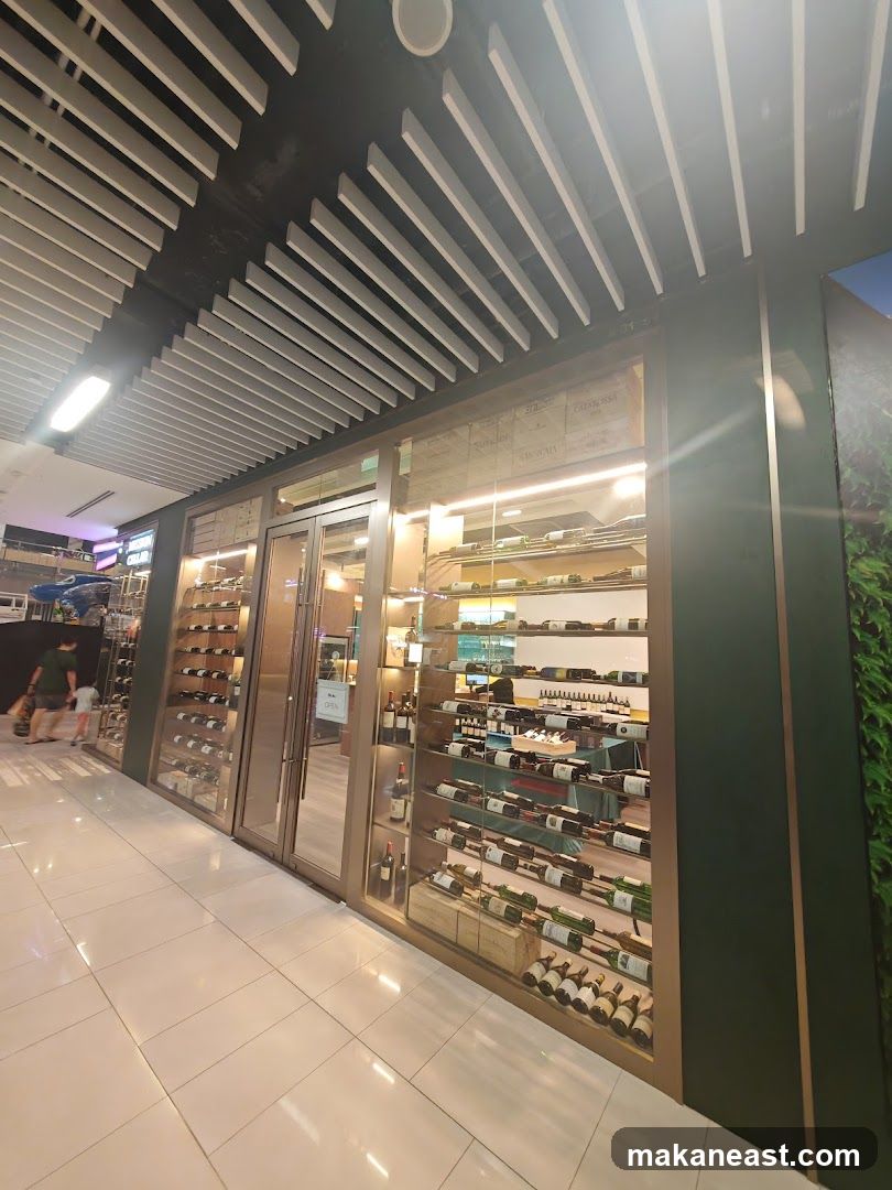 A well-lit wine store with a modern facade. The entrance features double glass doors, and the interior is visible through large glass windows showcasing numerous bottles of wine displayed on shelves. The shelves are stocked with various types of wine bottles, some in wooden crates. The store appears to be located within a mall or indoor shopping area, with a tiled floor and a distinctive slatted ceiling. People are seen walking by outside the store.