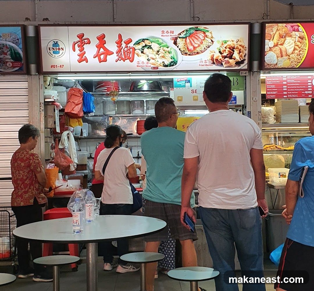 A bustling food stall with a prominent sign displaying "Union Wanton Noodle" in Chinese and English, along with images of their dishes. The stall is busy with customers queuing and staff preparing food. The storefront features a counter with food preparation areas and shelves stocked with ingredients and dishes.