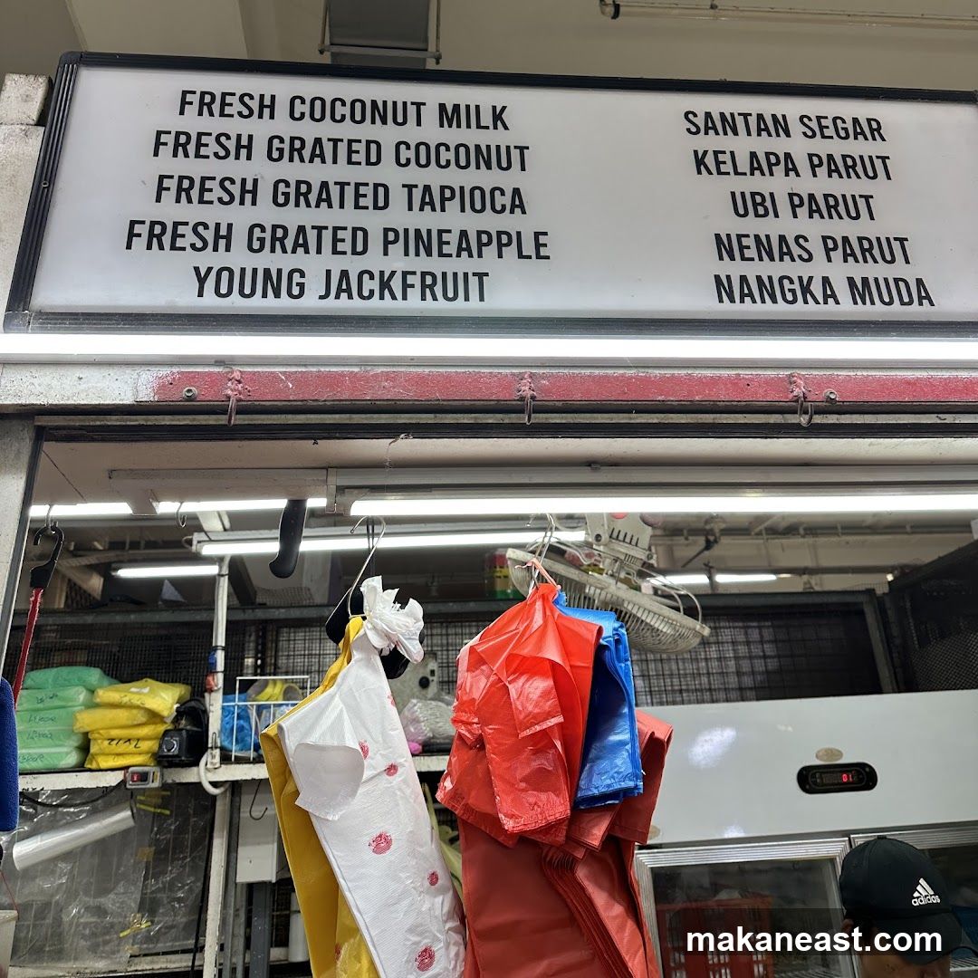 A menu board lists fresh coconut milk and various grated items in English and Malay. Below the menu, plastic bags hang, and a person wearing an Adidas cap is partially visible.