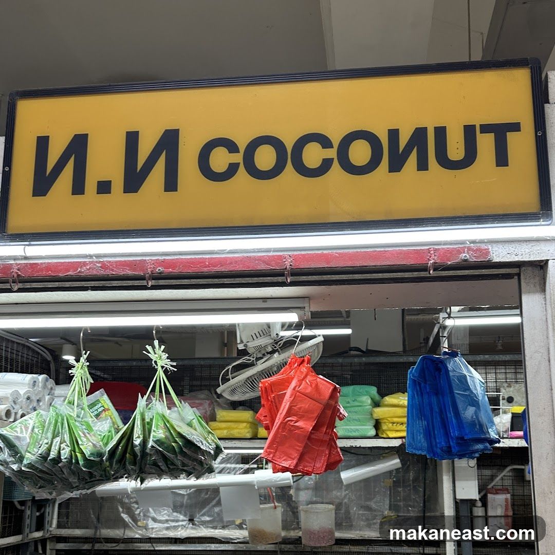 A yellow sign with black lettering reads "N.N COCONUT" above a stall selling various items. Green, red, and blue bags are hanging, along with rolls of plastic wrap. Shelves behind the hanging items display stacks of yellow and green items. A fan is visible in the background.