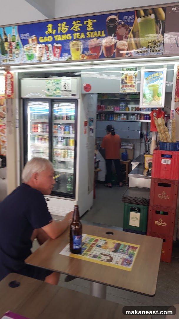 The exterior of a tea stall with a large sign displaying drinks and the stall name. A man sits at a table in the foreground with a bottle of beer. Inside, shelves are stocked with various items, and a person is working behind the counter. Refrigerated display cases are visible, filled with beverages.