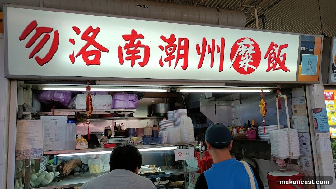A hawker stall with a large illuminated sign in red Chinese characters. Below the sign, the stall counter is visible with stacks of plates and containers. Two customers are standing in front of the counter.