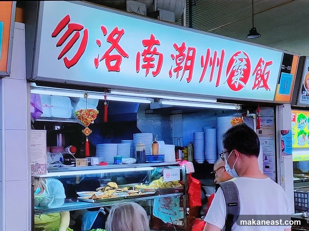 A brightly lit food stall with a large sign in red Chinese characters. Below the sign, food items are displayed in a glass counter, and stacks of plates are visible. People are seen queuing or ordering at the stall.
