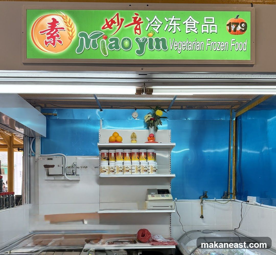 A brightly lit stall with a prominent green sign above that reads "Mao Yin Vegetarian Frozen Food" in English and Chinese characters. The sign also features a wheat stalk and a pumpkin graphic. Below the sign, shelves display various packaged food items, oranges, and decorative items. A cash register is visible on a shelf, and a glass-covered freezer unit is in the foreground. The stall has a blue tiled back wall.