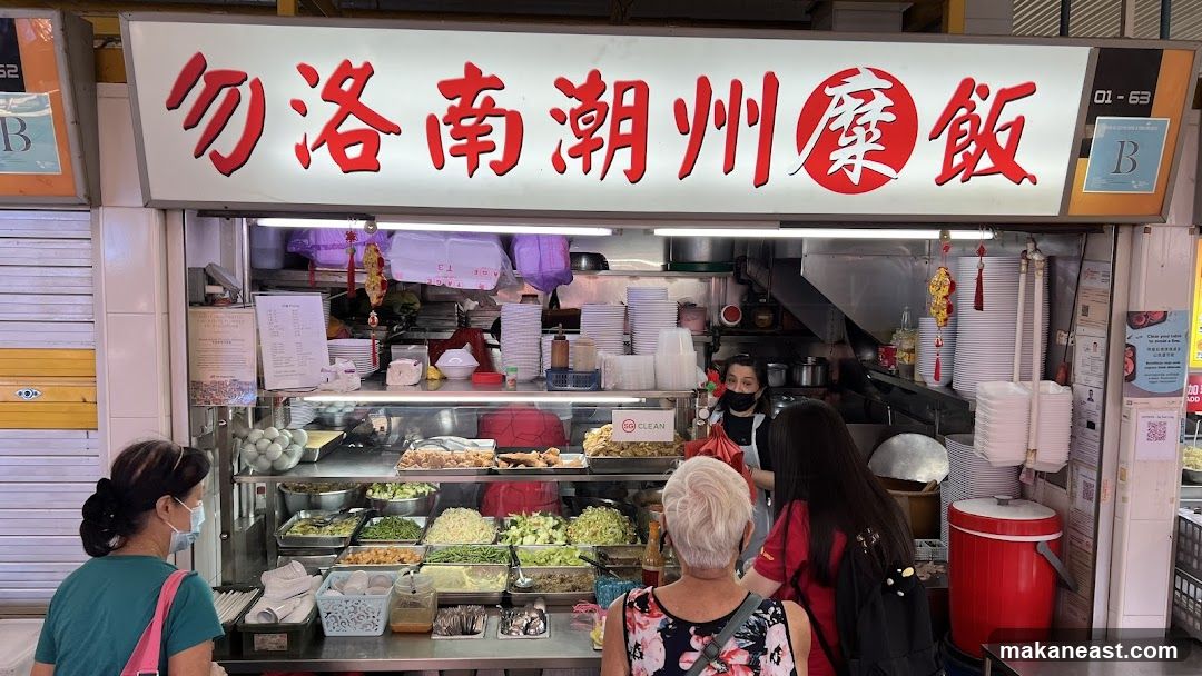 A food stall with a prominent illuminated sign in Chinese characters. Below the sign, a display case showcases various dishes, including what appear to be eggs, vegetables, and fried items. Several people are visible, some ordering food and others working behind the counter. Stacks of plates and containers are also present.