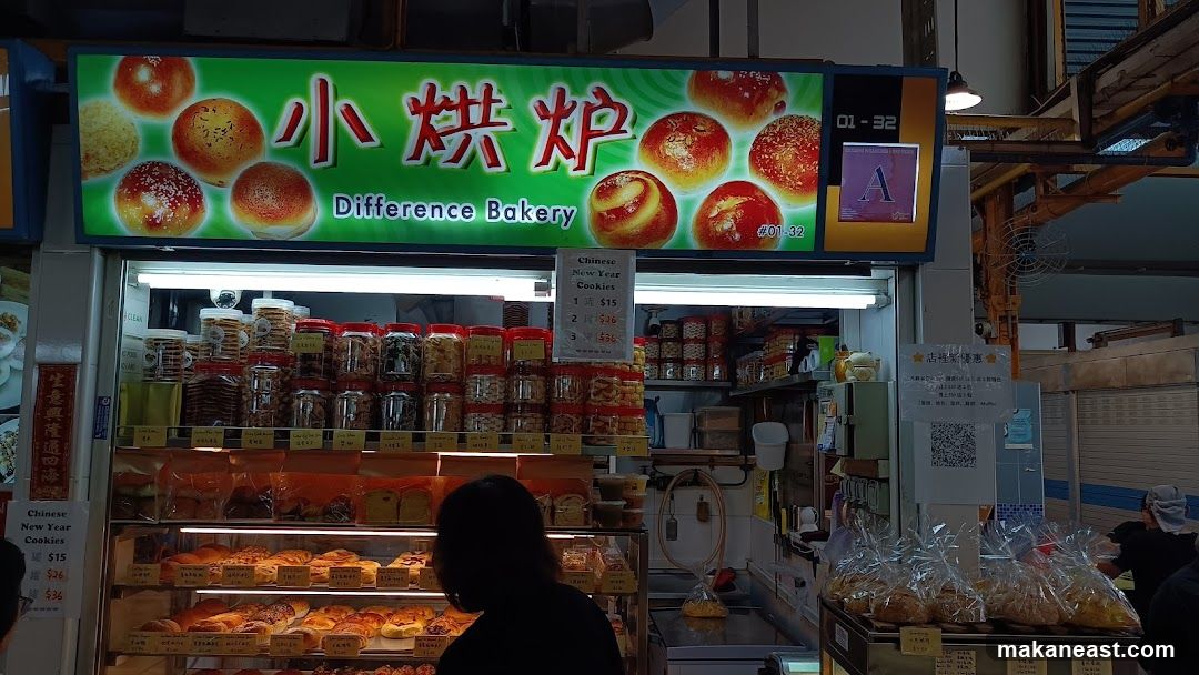 A bakery storefront with a bright green sign featuring images of buns and the text "小烘炉 Difference Bakery". Below the sign, shelves are stocked with jars of cookies and a display case filled with various baked goods. A price list for "Chinese New Year Cookies" is visible.