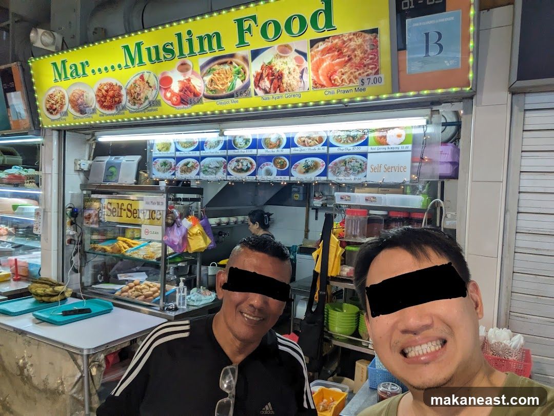 A food stall with a large yellow sign that reads "Mar.....Muslim Food" and displays pictures of various dishes. Below the sign is a menu board with prices and dish names. Two men are posing for a photo in front of the stall. The stall appears to be a self-service establishment.