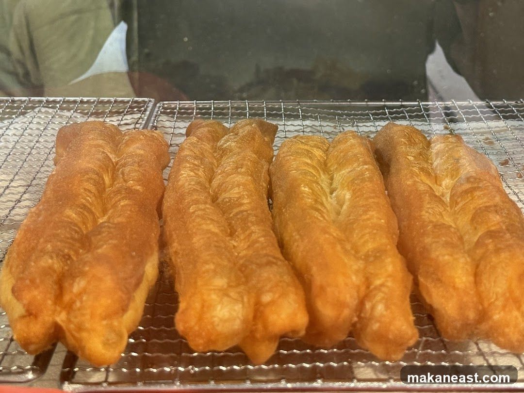 Four golden-brown fried dough sticks are arranged side-by-side on a metal cooling rack. The dough sticks have a distinctive twisted appearance, with two prominent ridges running along their length. The background is blurred, showing a dark reflective surface, possibly a glass display case or window.