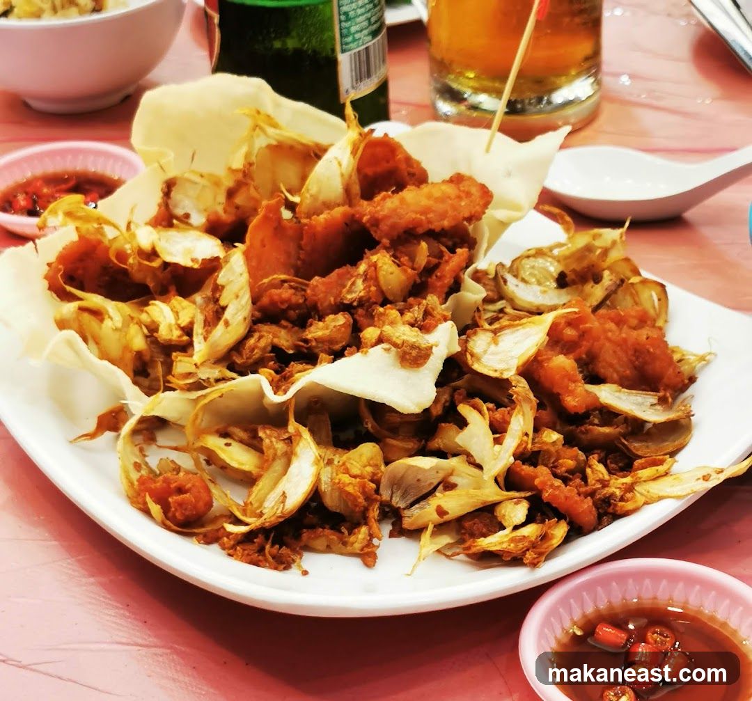 A close-up shot of a dish featuring fried garlic, crispy pork pieces, and what appear to be fried onion slices, served in a white bowl lined with a light-colored crisp. Two small pink bowls containing a dipping sauce with chili slices are visible on the sides. In the background, a green bottle and a glass of amber liquid, possibly beer, are present. The table surface is pink.