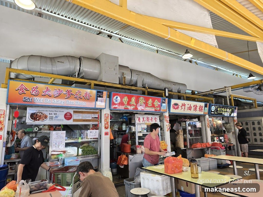 A bustling food stall in a market setting, with various vendors and customers. The stall in the foreground is named 'Shi Wei Da Satay Bee Hoon' and offers satay bee hoon for $4/$5. Other stalls visible include '香美斋素食' (Xiang Mei Zhai Vegetarian Food) and a stall selling fried dough sticks. People are seen preparing and serving food, and customers are seated at tables. The overall atmosphere is lively and typical of a hawker center.