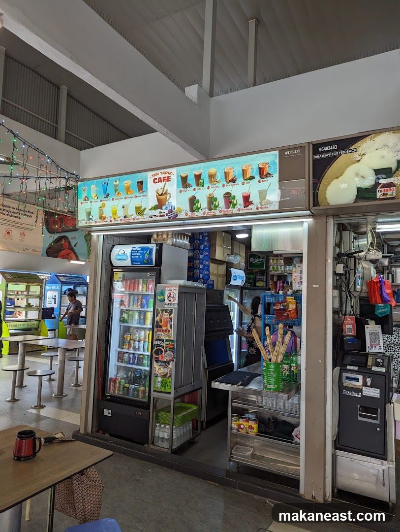 A stall with a large illuminated menu board displaying various drinks and food items. A refrigerator stocked with beverages is visible, along with shelves containing snacks and other products. The stall appears to be located in an open-air food court or market.
