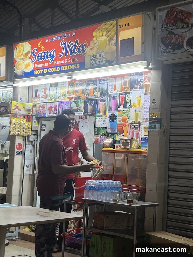 A brightly lit storefront for a drink stall named Sang Nila, offering hot and cold drinks. The sign features images of drinks and the text "Minuman Panas & Sejuk" and "HOT & COLD DRINKS". Below the main sign, a menu board displays various drinks with prices. Two men are visible, one preparing a drink.