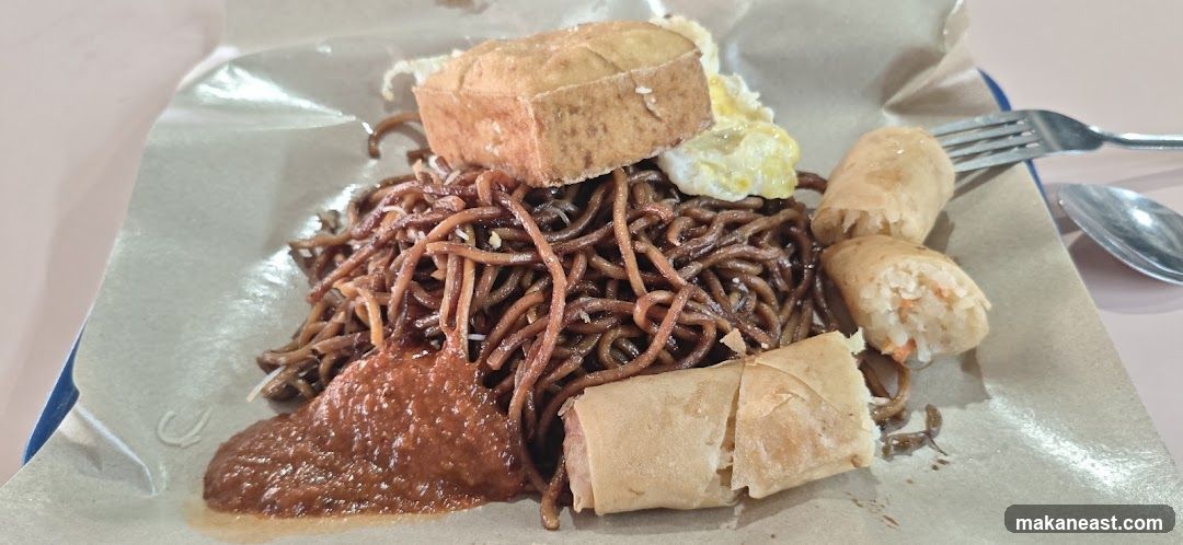 A plate of dark noodles is topped with a piece of fried tofu and a fried egg. Three spring rolls are arranged on the side. A dollop of red sauce is on the left. The food is served on brown paper. A fork and spoon are visible in the background.