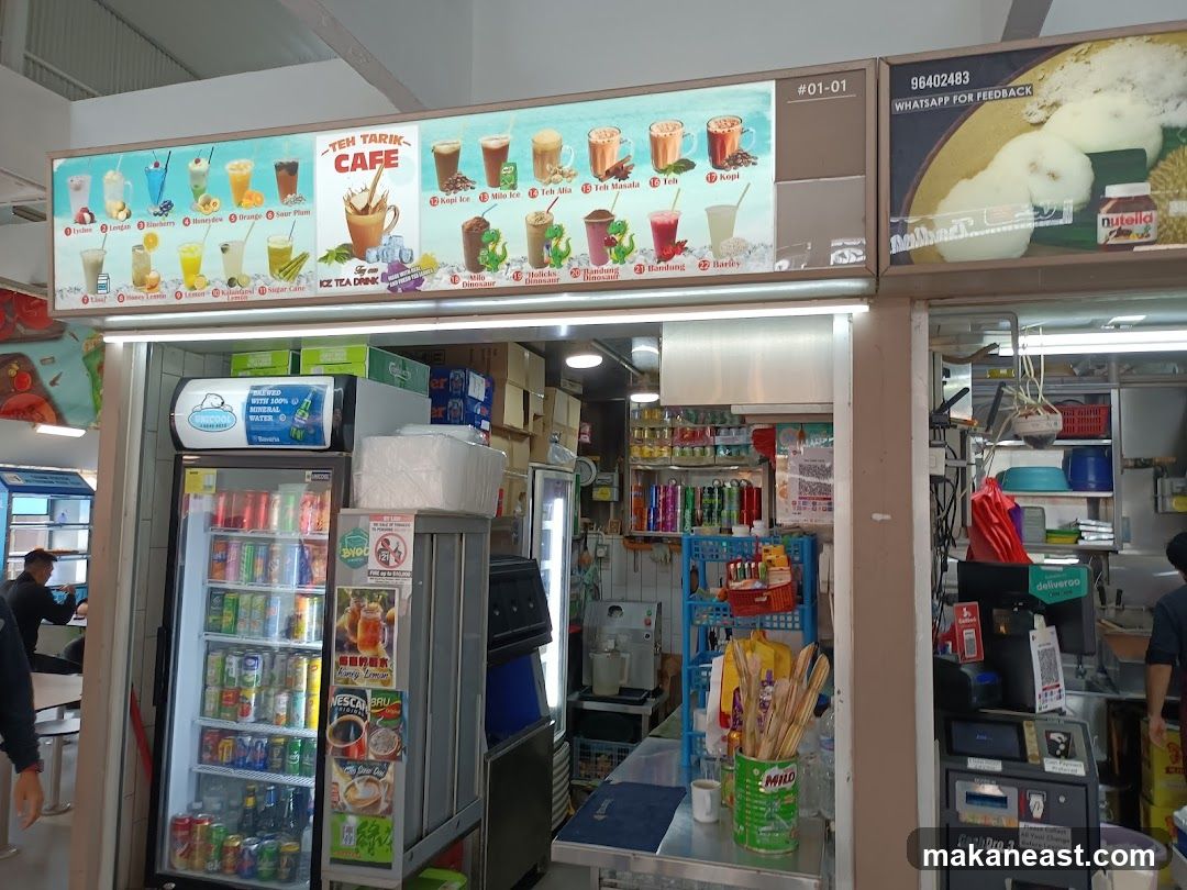 A brightly lit food stall with a large menu board displaying various drinks. The menu features illustrated drinks with names like Lychee, Lengan, Blueberry, Orange, Sour Plum, Kopi Ice, Milo Ice, Teh Alia, Teh Masala, Teh, Holicks, Bandung, and Barley. There are also images of durian and Nutella. Refrigerated drinks are visible in a cooler.