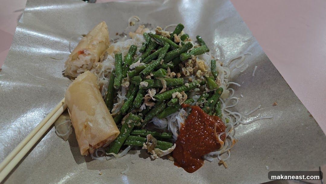 A close-up shot of a meal served on brown paper. The dish consists of thin white noodles topped with chopped green beans and scrambled egg. Two golden-brown spring rolls are placed on the side, and a dollop of red chili paste is visible. A pair of chopsticks rests on the paper.