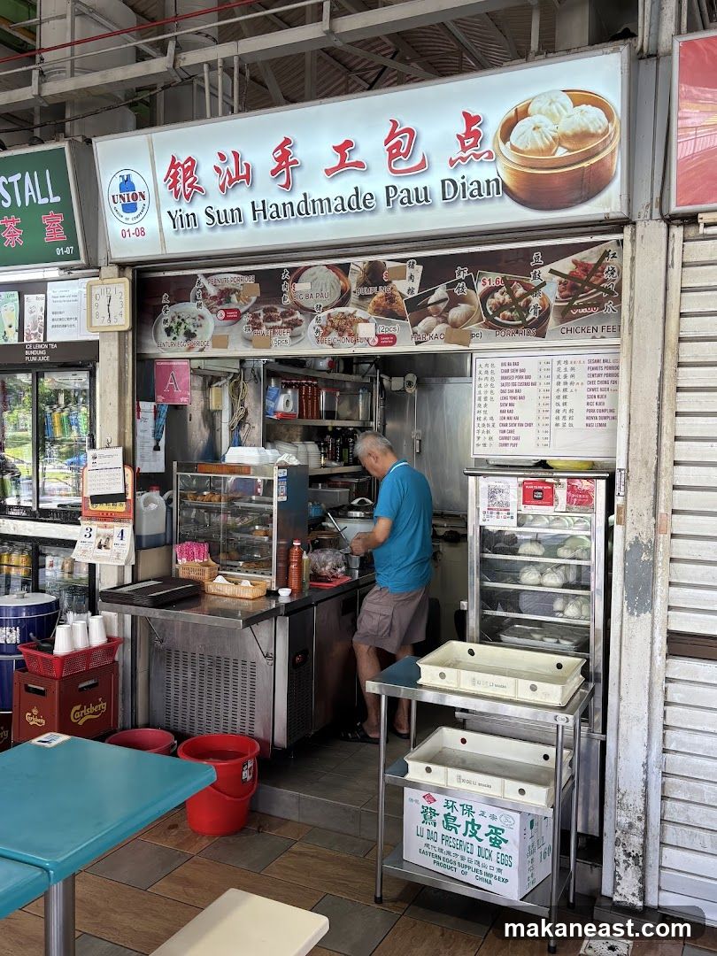 A street food stall with a sign that reads "Yin Sun Handmade Pau Dian" in English and Chinese. The stall displays various food items and has a menu board. A man is seen working behind the counter.